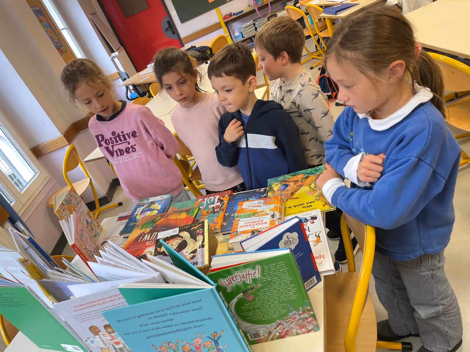 Vijf kinderen bekijken samen boeken op een tafel in een klaslokaal met gele stoelen.
