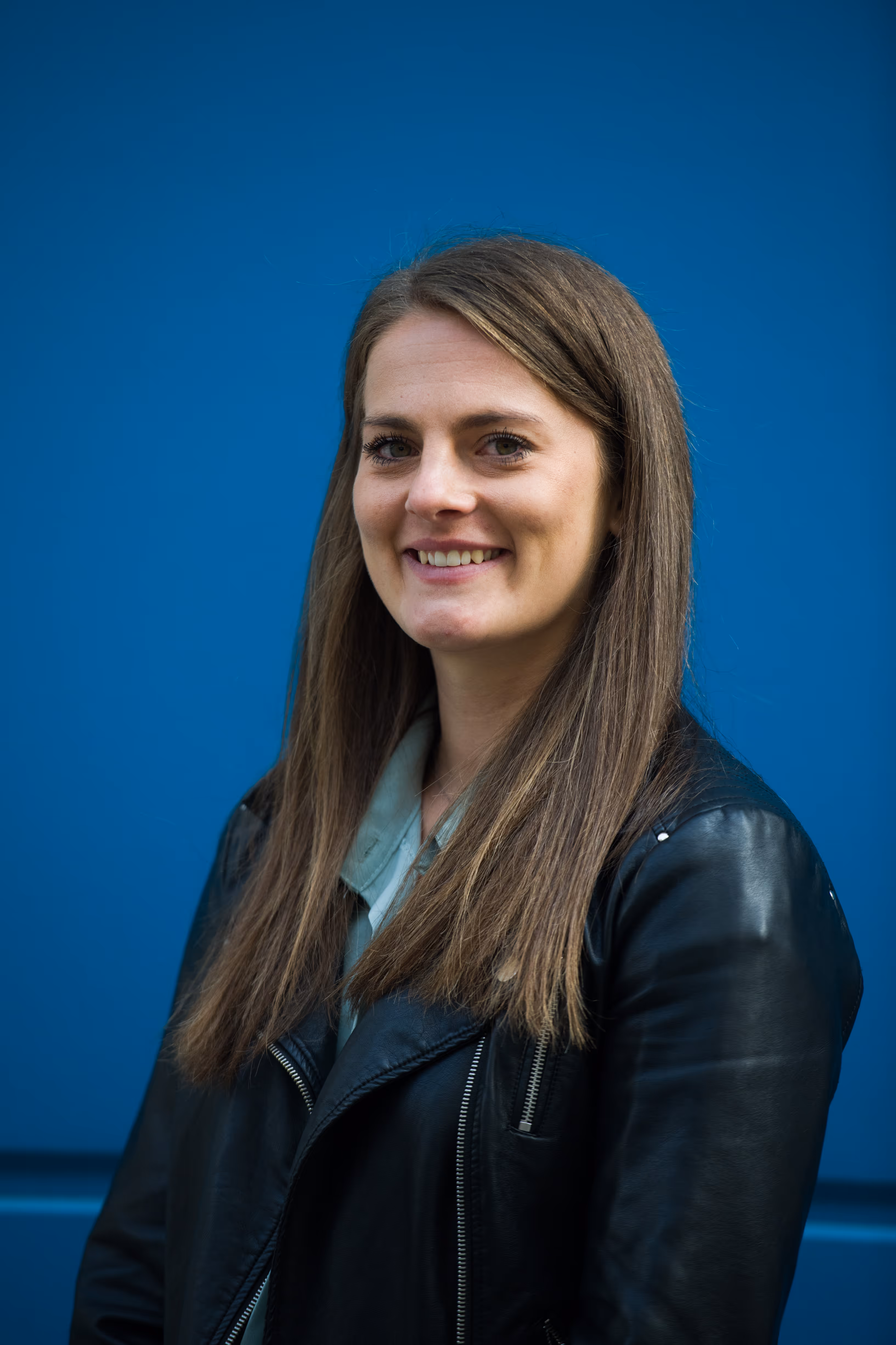 Vrouw met lang bruin haar draagt een zwarte leren jas en glimlacht voor een blauwe achtergrond.