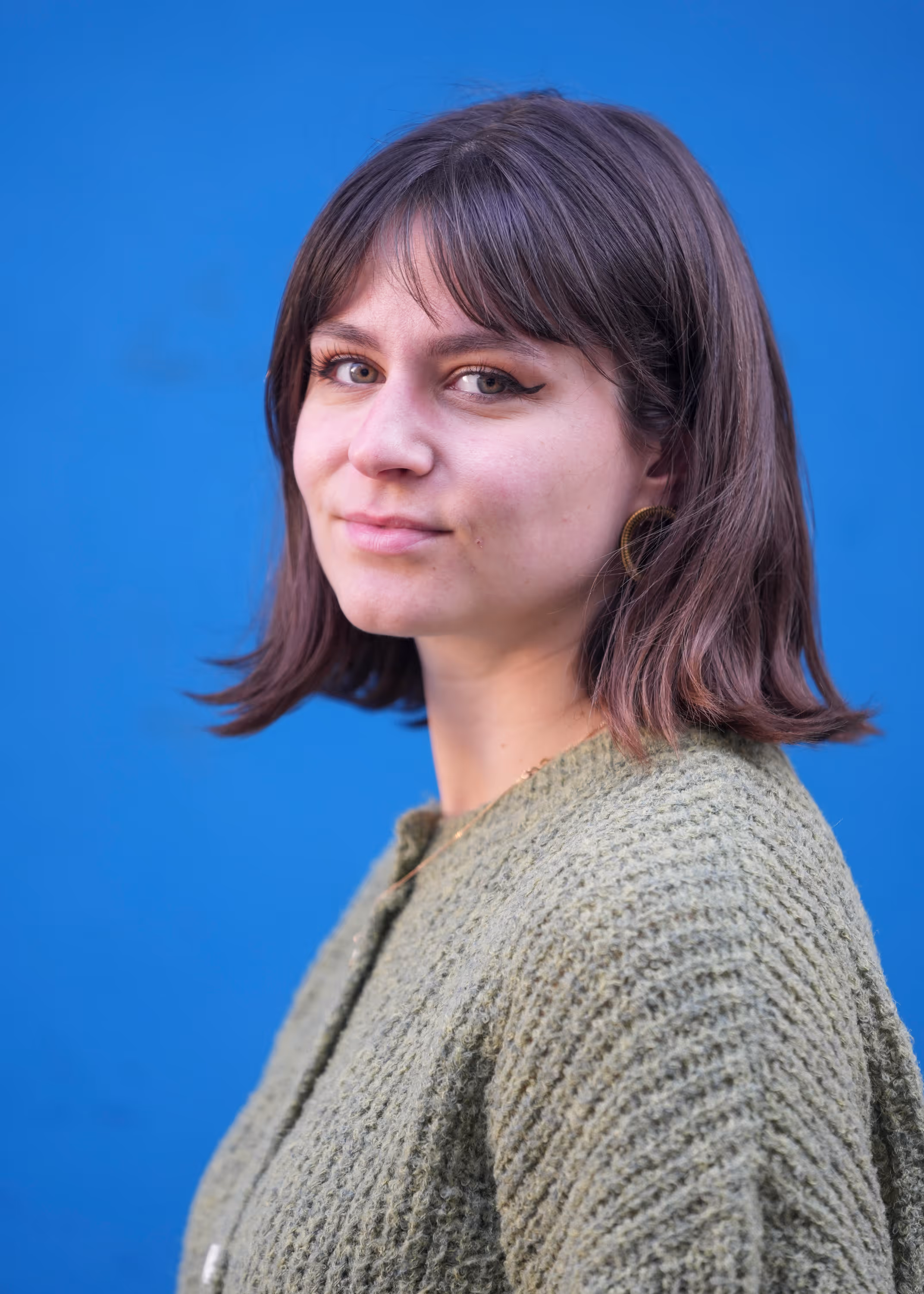 Jonge vrouw met halflang bruin haar draagt groene gebreide trui, kijkt naar de camera voor blauwe achtergrond.