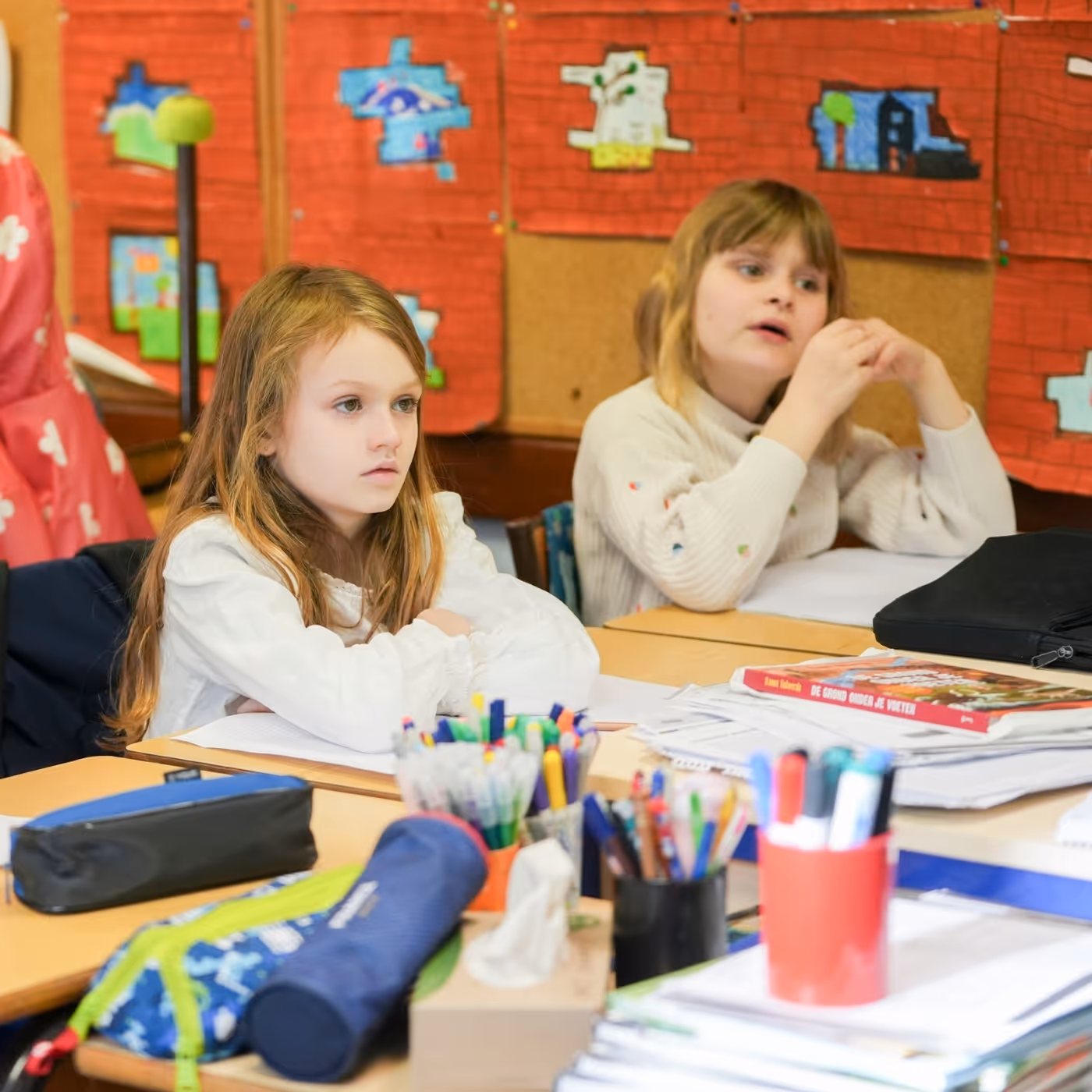 Twee meisjes zitten in de klas, omringd door pennen en schoolmateriaal op tafels.