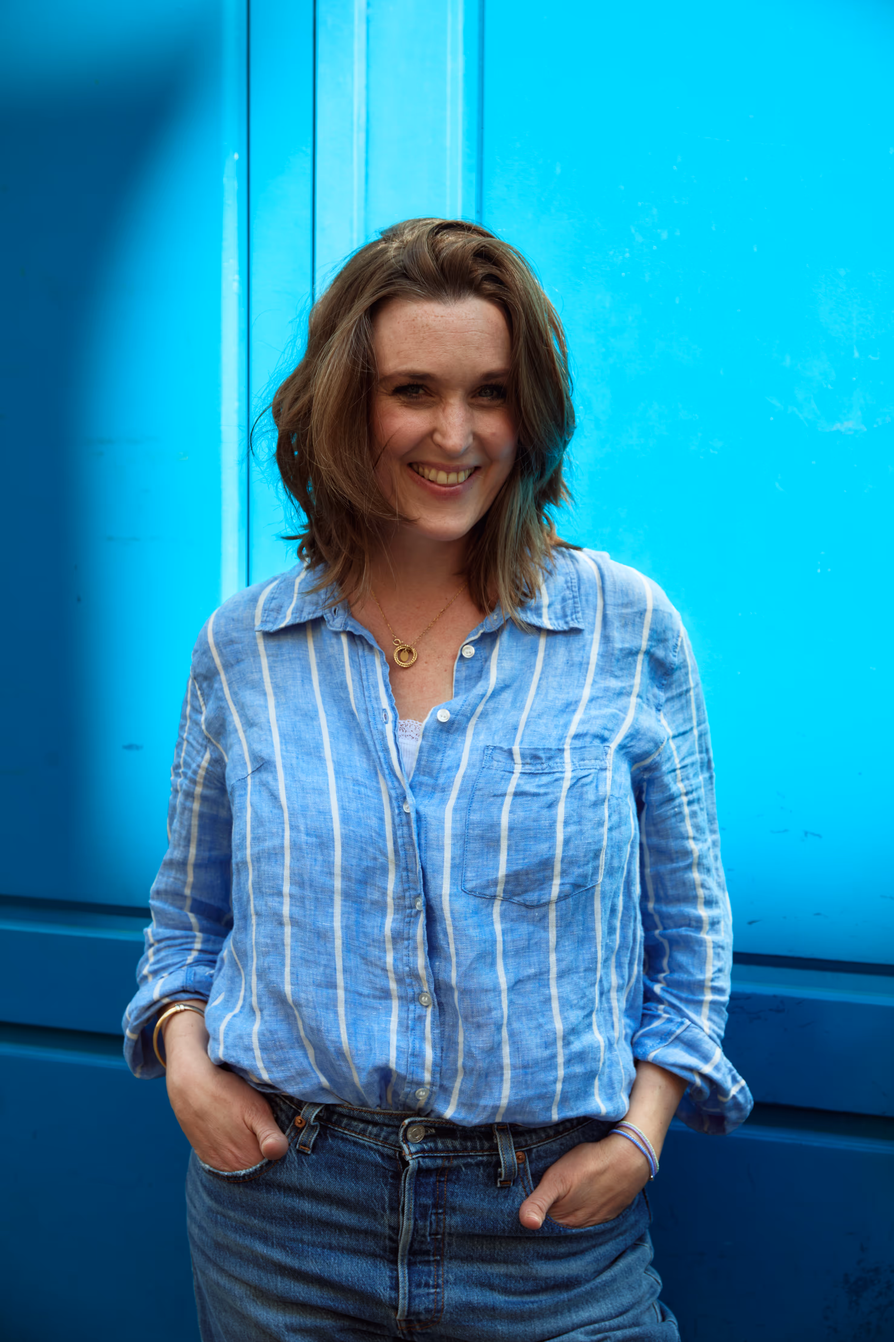 Vrouw met halflang haar lacht, draagt blauw gestreept shirt en spijkerbroek, handen in zakken tegen blauwe muur.