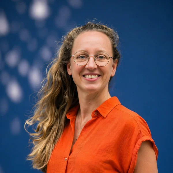 Vrouw met lang golvend haar en bril glimlacht, draagt oranje blouse voor blauwe achtergrond.