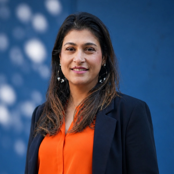 Vrouw met lang donker haar in oranje blouse en zwarte blazer voor blauwe achtergrond met lichtvlekken.