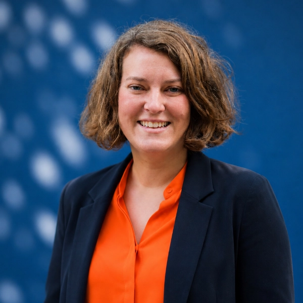Vrouw met kort golvend haar lacht, draagt oranje blouse en donker jasje tegen blauwe achtergrond.
