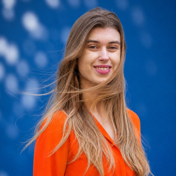 Jonge vrouw met lang haar en oranje blouse, glimlachend voor een blauwe achtergrond.