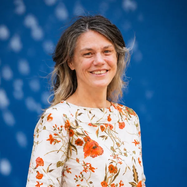 Vrouw met halflang haar lacht, ze draagt een wit shirt met oranje bloemen en staat voor een blauwe achtergrond.