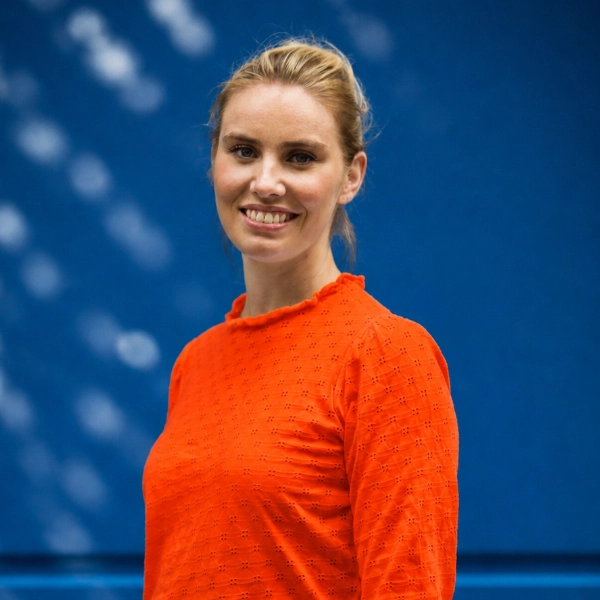 Vrouw met blond haar in een oranje trui lachend voor een blauwe achtergrond.