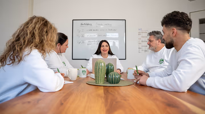 Fünf Personen in weißen Pullovern sitzen um einen Holztisch in einem Besprechungsraum mit einem Laptop und Kaffeetassen.