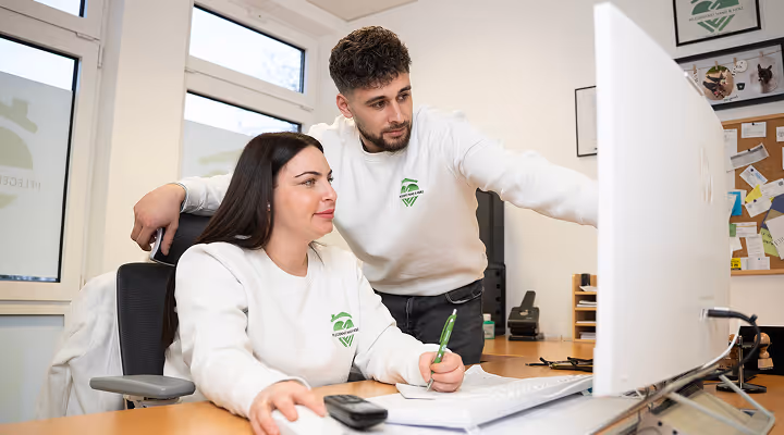 Zwei Kollegen im Büro sitzen vor einem Computerbildschirm, der Mann zeigt etwas darauf, während die Frau einen grünen Stift hält.