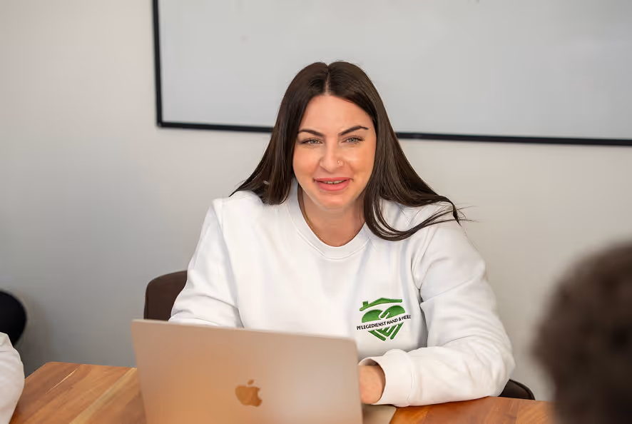 Frau mit dunklen Haaren und weißem Pullover mit grünem Pflegedienst-Logo sitzt lächelnd vor einem Laptop in einem Besprechungsraum.