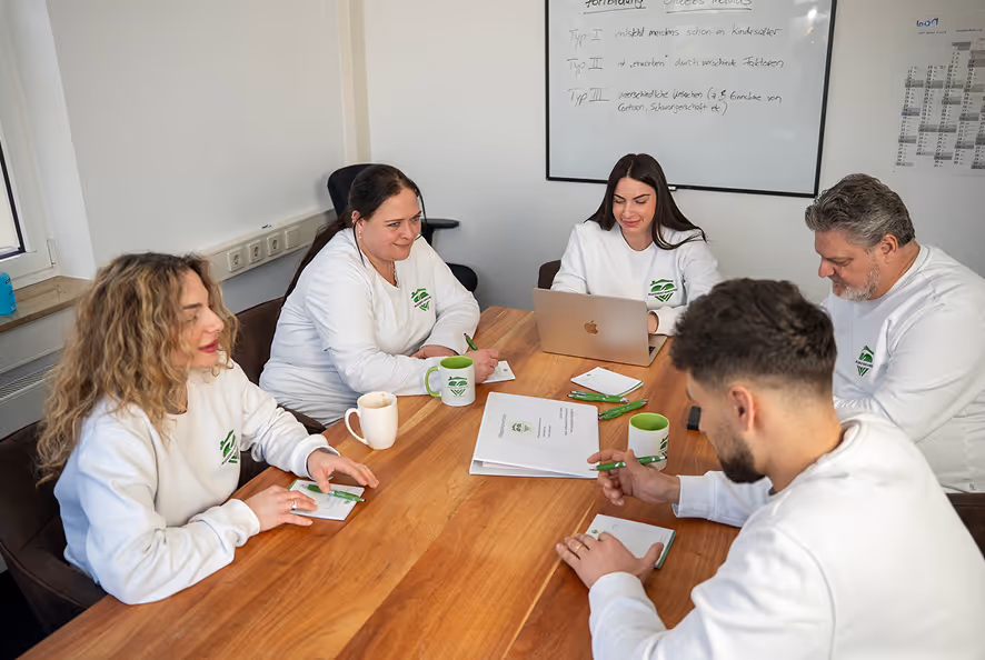 Fünf Personen in weißen Pullovern sitzen an einem Tisch in einem Besprechungsraum und führen eine Teamdiskussion.