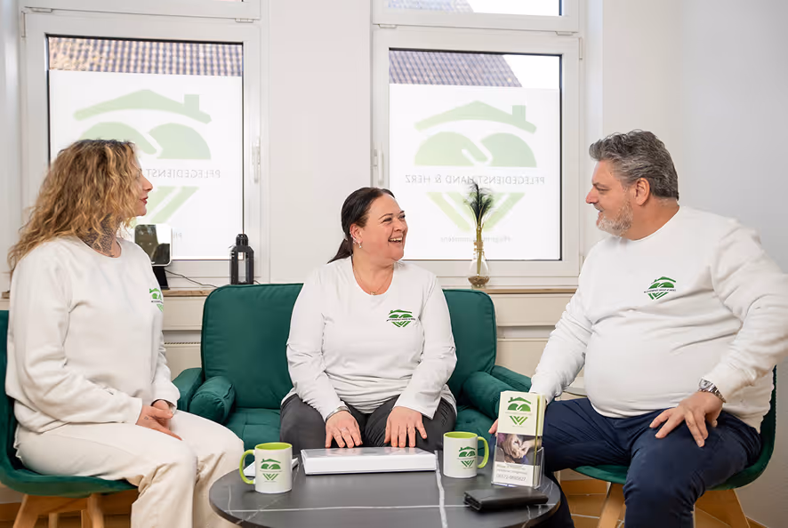 Drei Personen in weißen Pullovern mit grünem Logo sitzen um einen runden Tisch und unterhalten sich in einem hellen Raum.