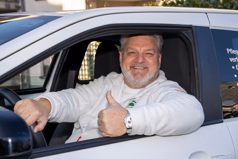 Mann in weißem Pullover sitzt in einem weißen Auto, lächelt und zeigt Daumen hoch.