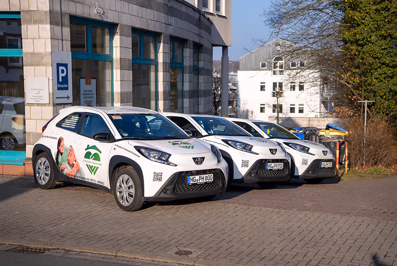 Drei weiße Autos mit Werbeaufklebern für einen ambulanten Pflegedienst parken nebeneinander vor einem Gebäude.