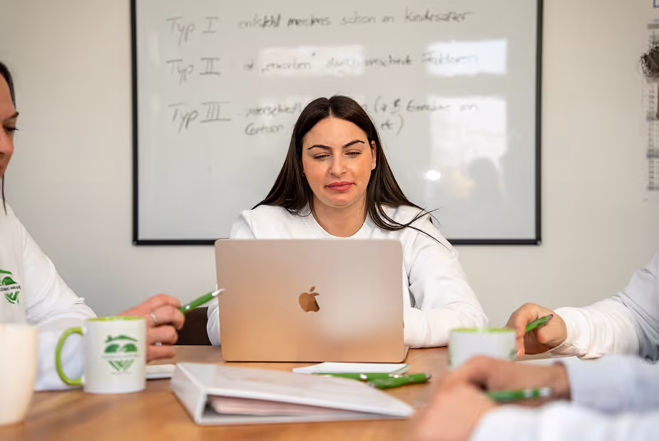 Frau sitzt an einem Tisch und arbeitet an einem Laptop vor einem Whiteboard, zwei weitere Personen sitzen daneben.