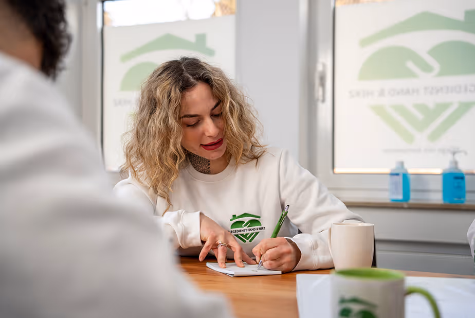 Frau mit lockigem Haar sitzt an einem Tisch und schreibt in ein Notizbuch.