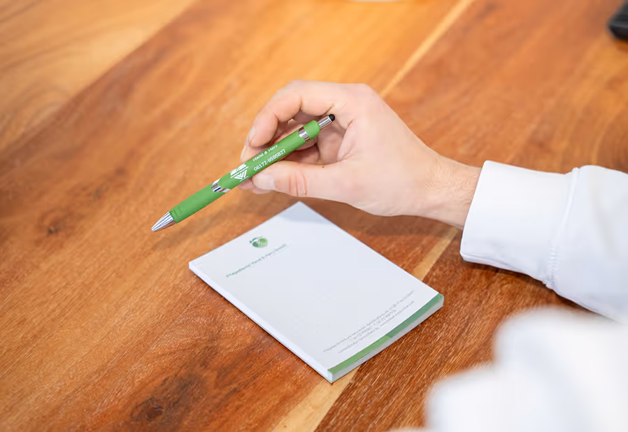 Eine Hand hält einen grünen Kugelschreiber über einem weißen Notizblock auf einem Holztisch.