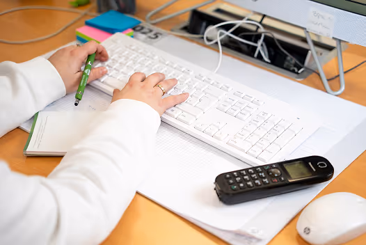 Person tippt auf einer weißen Tastatur und hält einen grünen Stift über einem Notizbuch auf einem Schreibtisch mit Telefon und Computermaus.