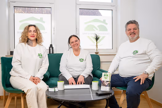 Drei lächelnde Menschen in weißen Pullovern mit grünem Logo sitzen an einem runden Tisch mit Tassen, im hellen Raum mit Fenstern im Hintergrund.