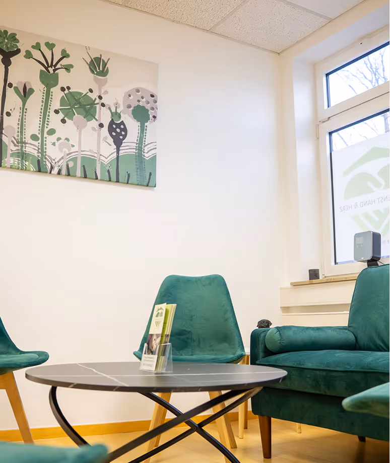 Wartebereich mit grünen Stühlen, rundem schwarzen Tisch und einem Gemälde mit grünen Pflanzendesigns an der Wand.