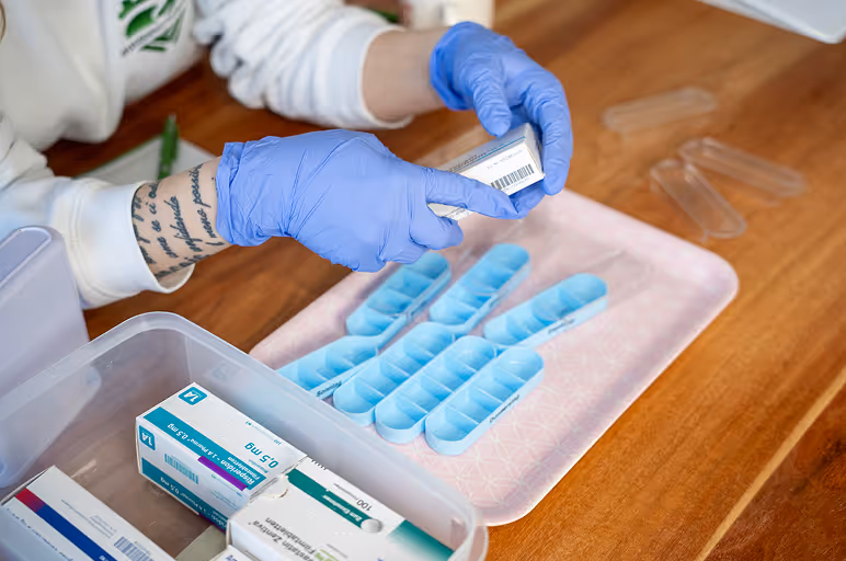 Eine Person mit blauen Handschuhen verteilt Medikamente aus einer Packung in blaue Pillendosen auf einem Tablett.
