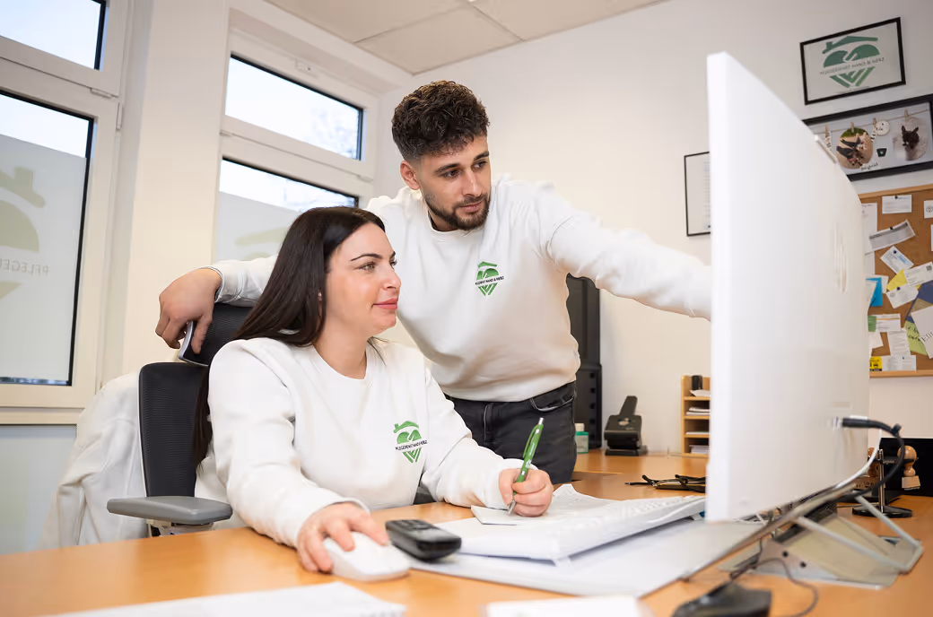 Eine Frau sitzt an einem Schreibtisch und benutzt eine Computermaus, während ein Mann neben ihr steht und auf den Monitor zeigt.