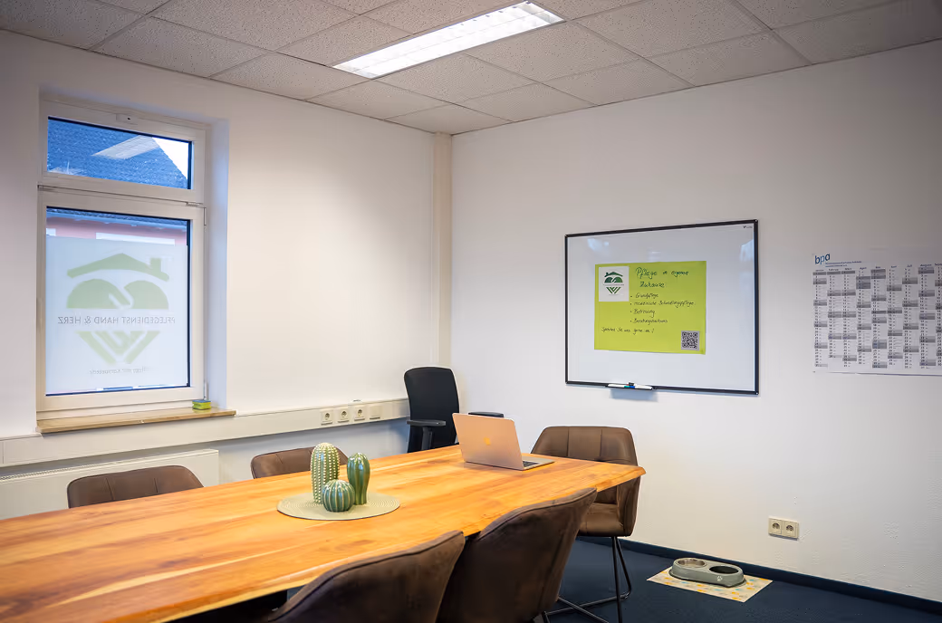 Konferenzraum mit Holztisch, mehreren braunen Stühlen, einem Laptop, einer weißen Tafel mit einem grünen Poster, und Futternäpfen auf dem Boden.