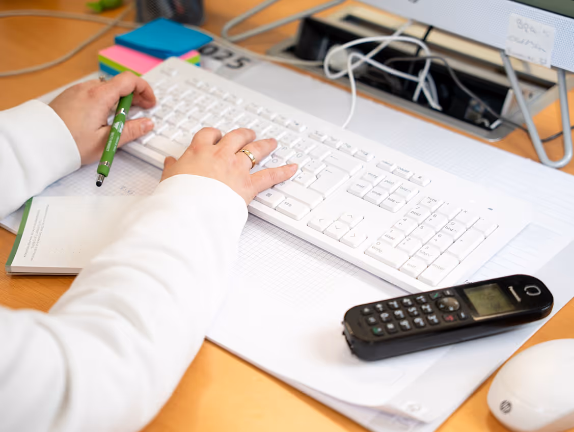 Person tippt auf einer weißen Tastatur und hält einen grünen Stift auf einem Schreibtisch mit Telefon, Notizblöcken und einem Computermaus.