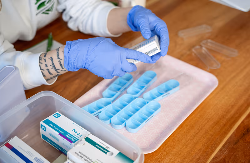 Person mit blauen Handschuhen hält eine Medikamentenpackung über Tablettenboxen auf einem Holztisch.