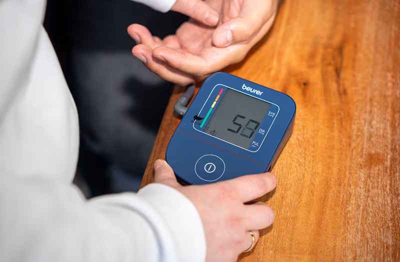 Eine Person misst ihren Blutdruck mit einem blauen Beurer Blutdruckmessgerät auf einem Holztisch.
