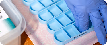 Hand mit blauen Handschuhen, die Tabletten aus einem blauen Tagesmedikamentenorganizer entnimmt.