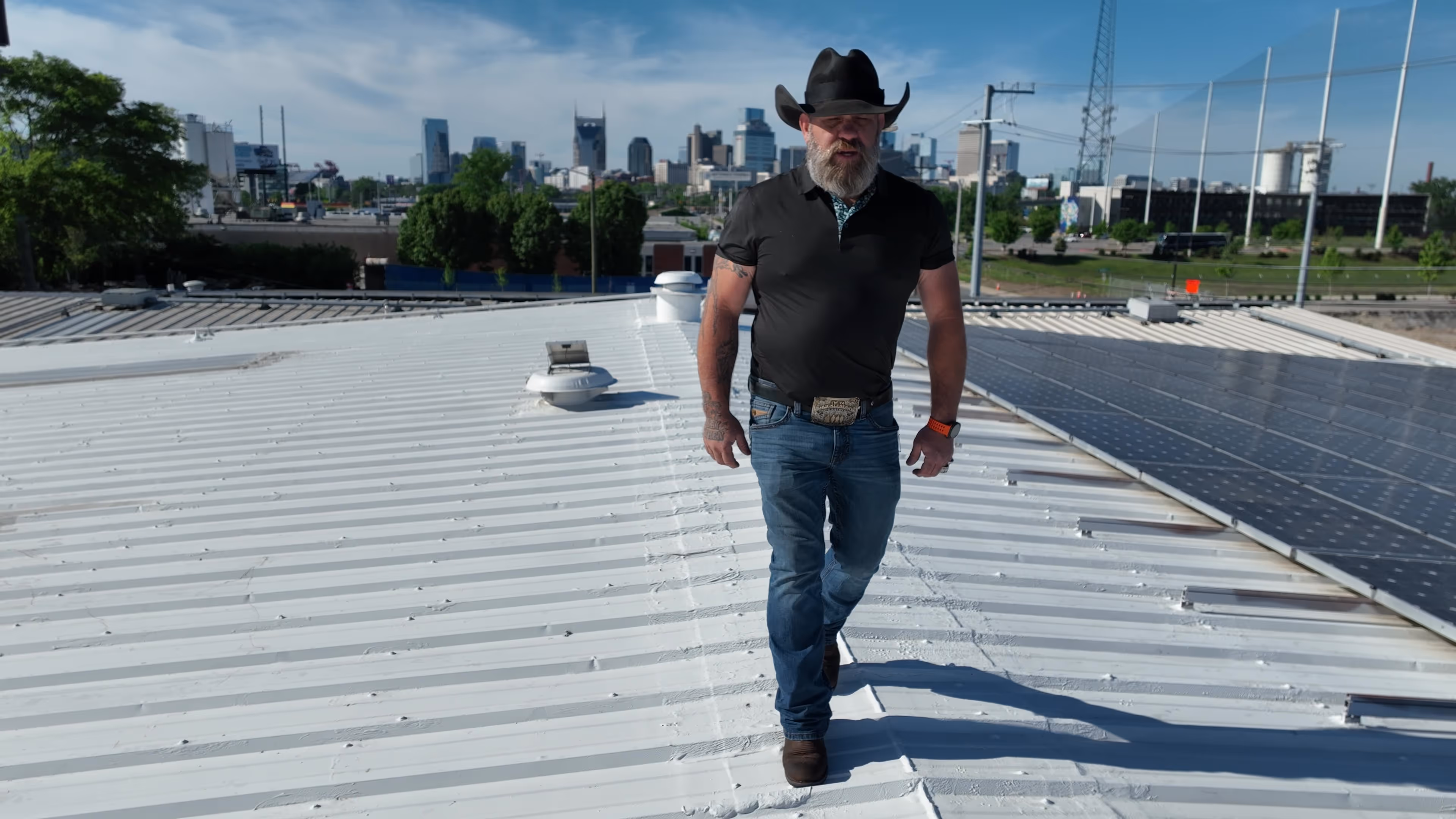 Bearded man in cowboy hat and jeans walking on a rooftop with solar panels and a city skyline behind.
