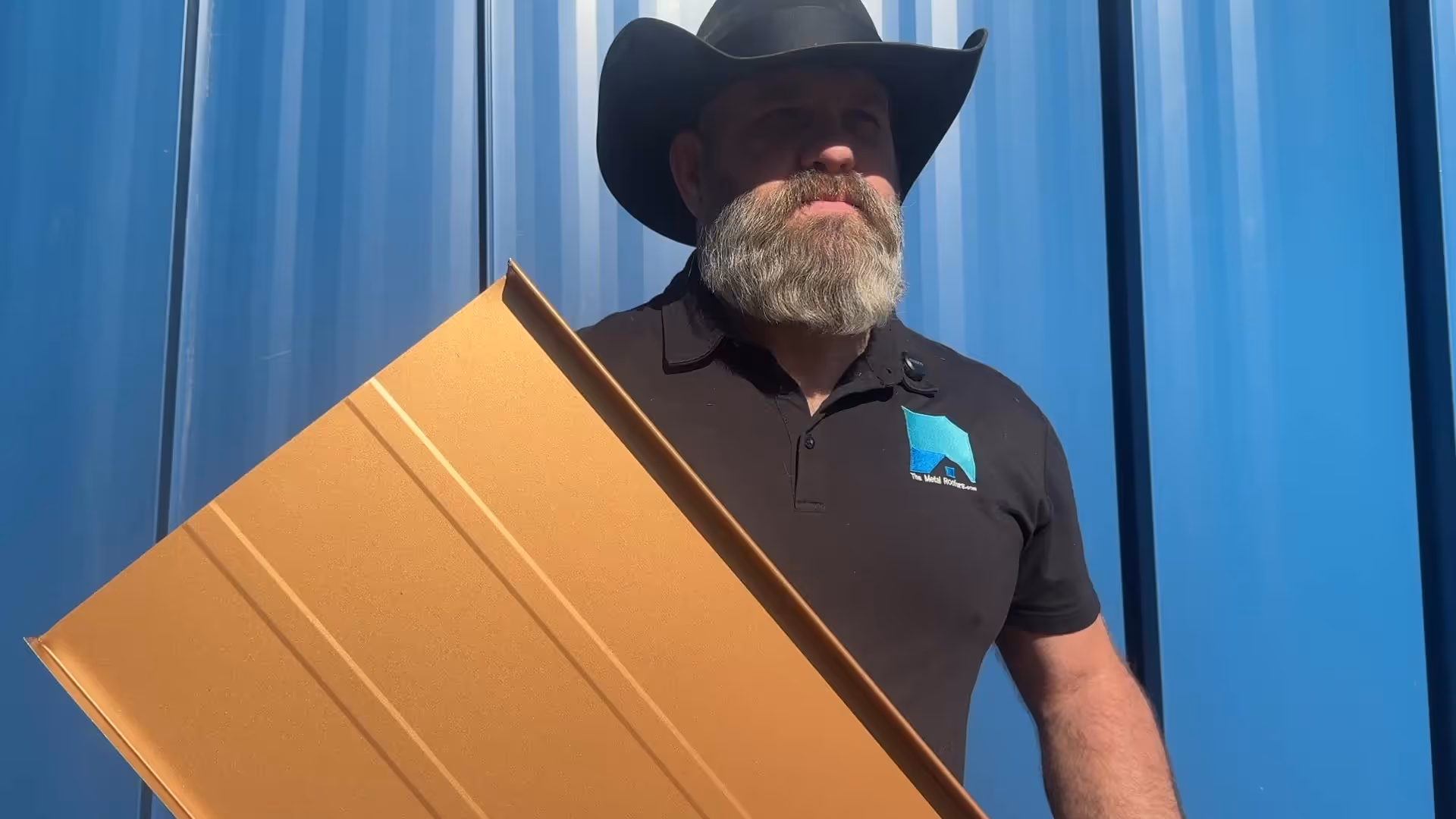 Bearded man in black cowboy hat holding a gold metal roofing panel in front of a blue wall.