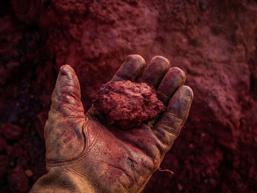 Worn leather-gloved hand holding a clump of red dirt over a red dirt ground.