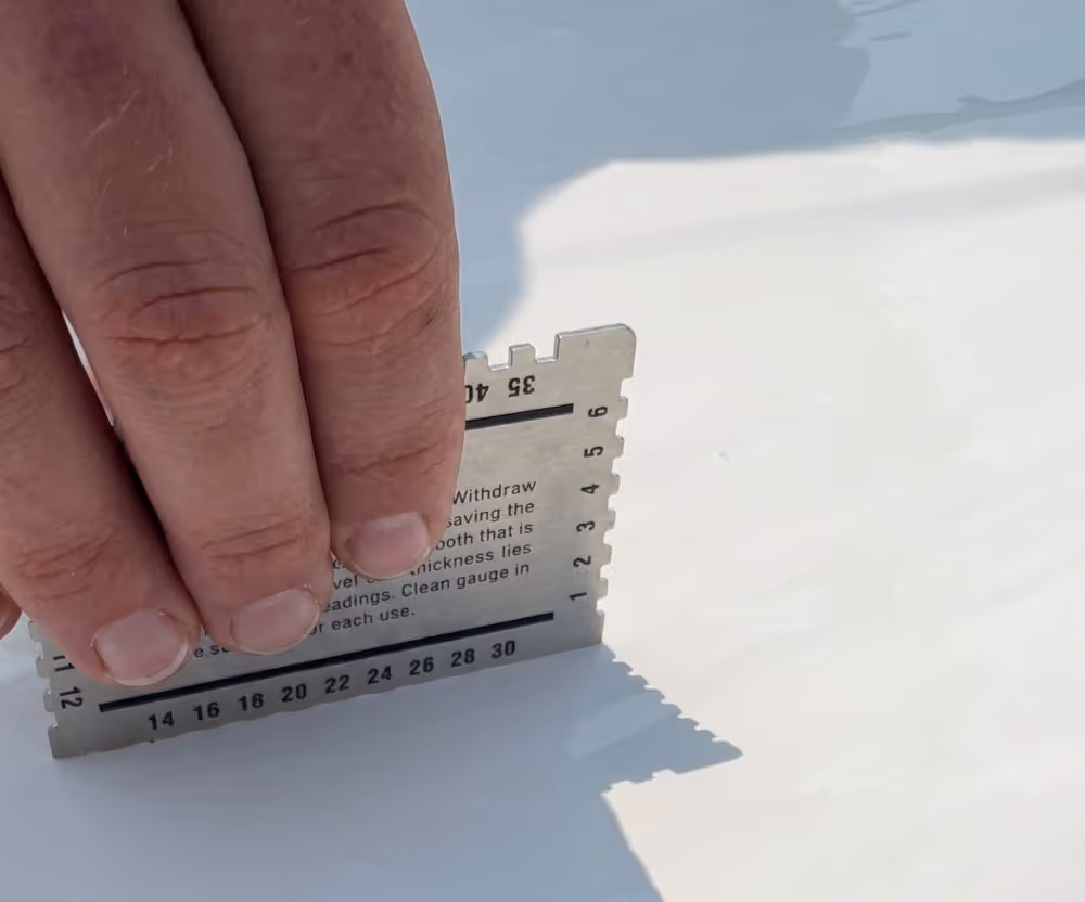 Hand holding a metal gauge tool on a white smooth surface to measure thickness.