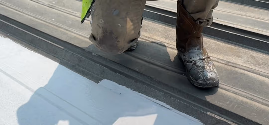 Worker wearing dirty boots kneeling on a metal roof being painted white on one side.
