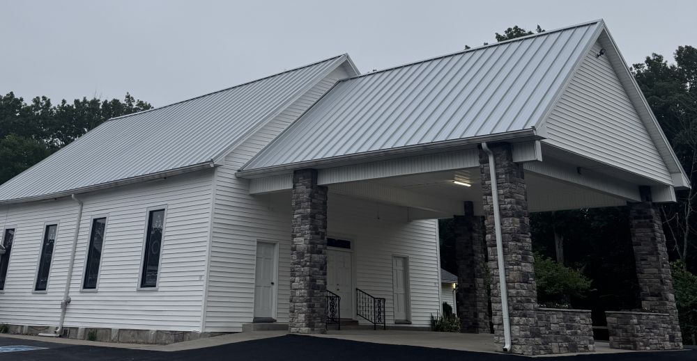 White building with metal roof and stone columns supporting a covered entrance on a paved area.