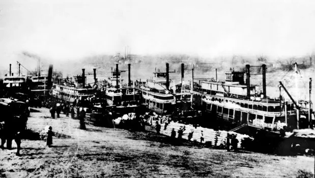 Black and white photo of several steamboats docked with people loading goods onshore.