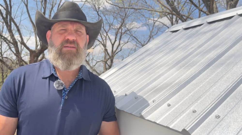 Man in a cowboy hat and blue shirt standing next to a silver metal roof with trees in the background.