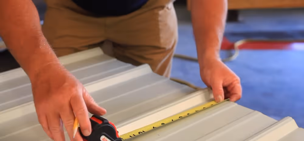 Person measuring a corrugated metal sheet with a tape measure and holding a pencil.