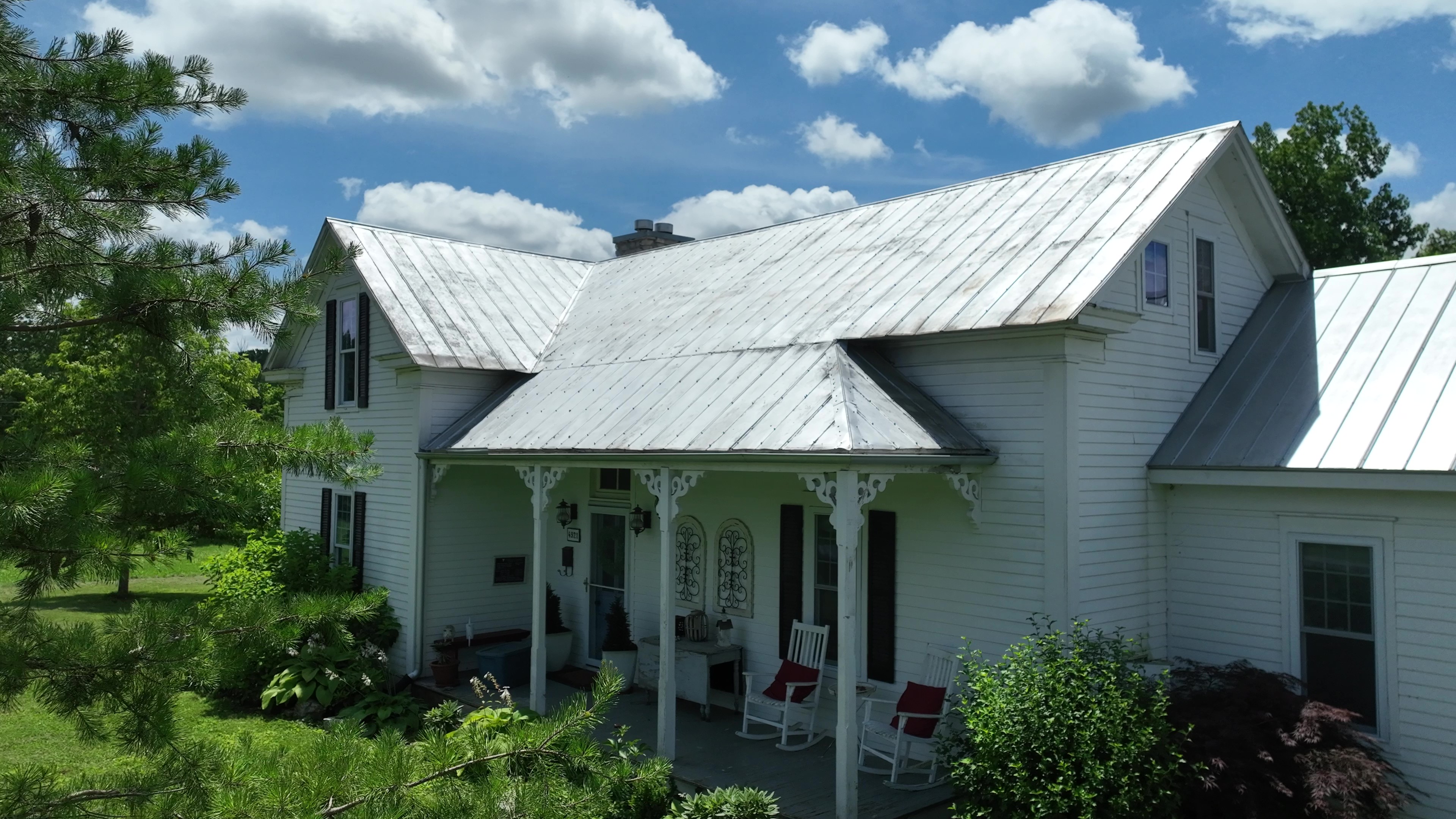 White house with metal roof, front porch with rocking chairs, surrounded by green trees and plants.