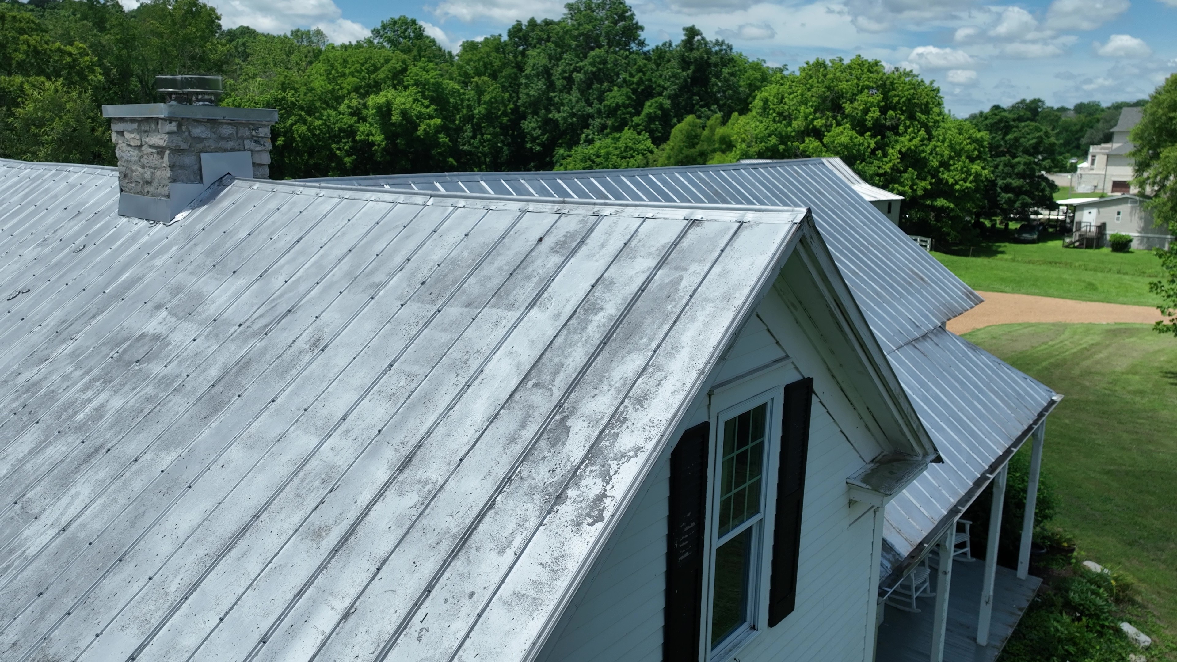White house with a weathered metal roof, chimney, and black shutters surrounded by green trees.