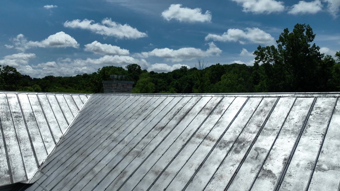 Silver metal roof with vertical ridges under a blue sky with scattered clouds and trees in the background.