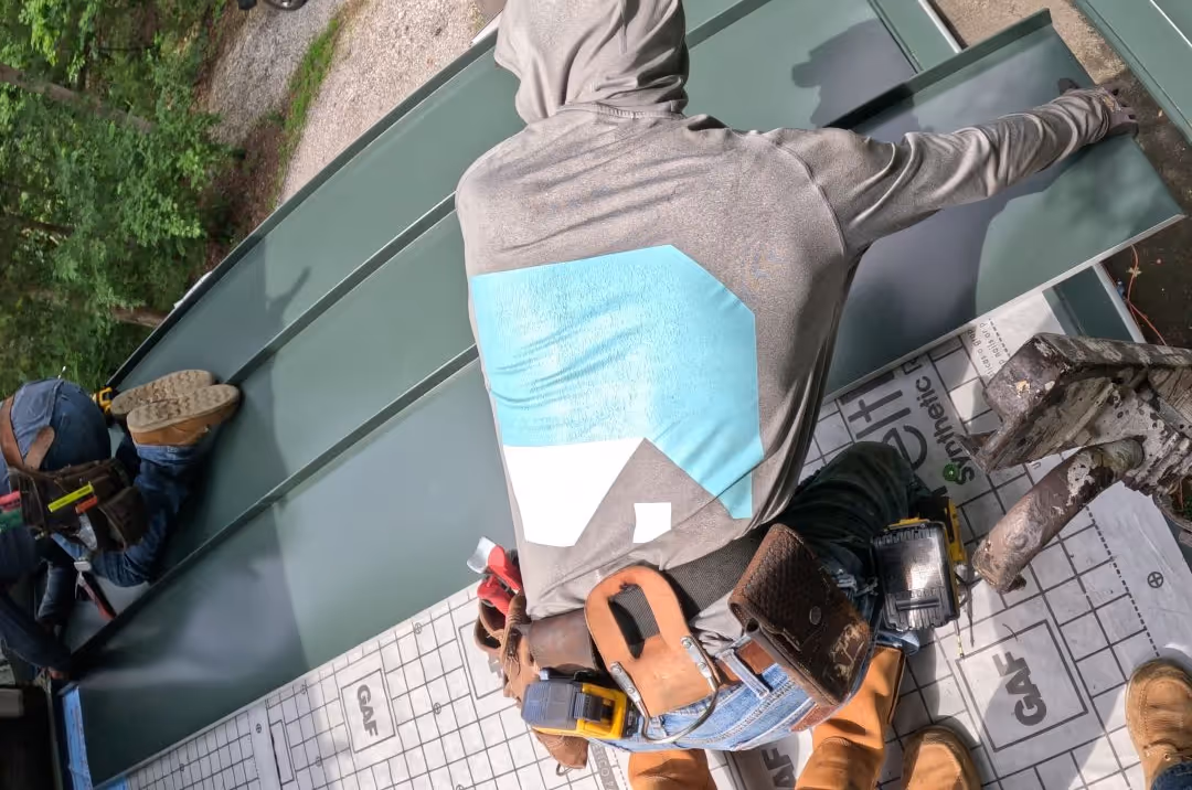 Two roofers in hoodies installing metal roof panels on a wooden roof base covered with GAF synthetic underlayment.