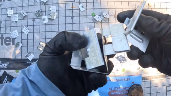 Hands in black gloves holding metal mounting brackets over a grid-patterned surface with scattered hardware.