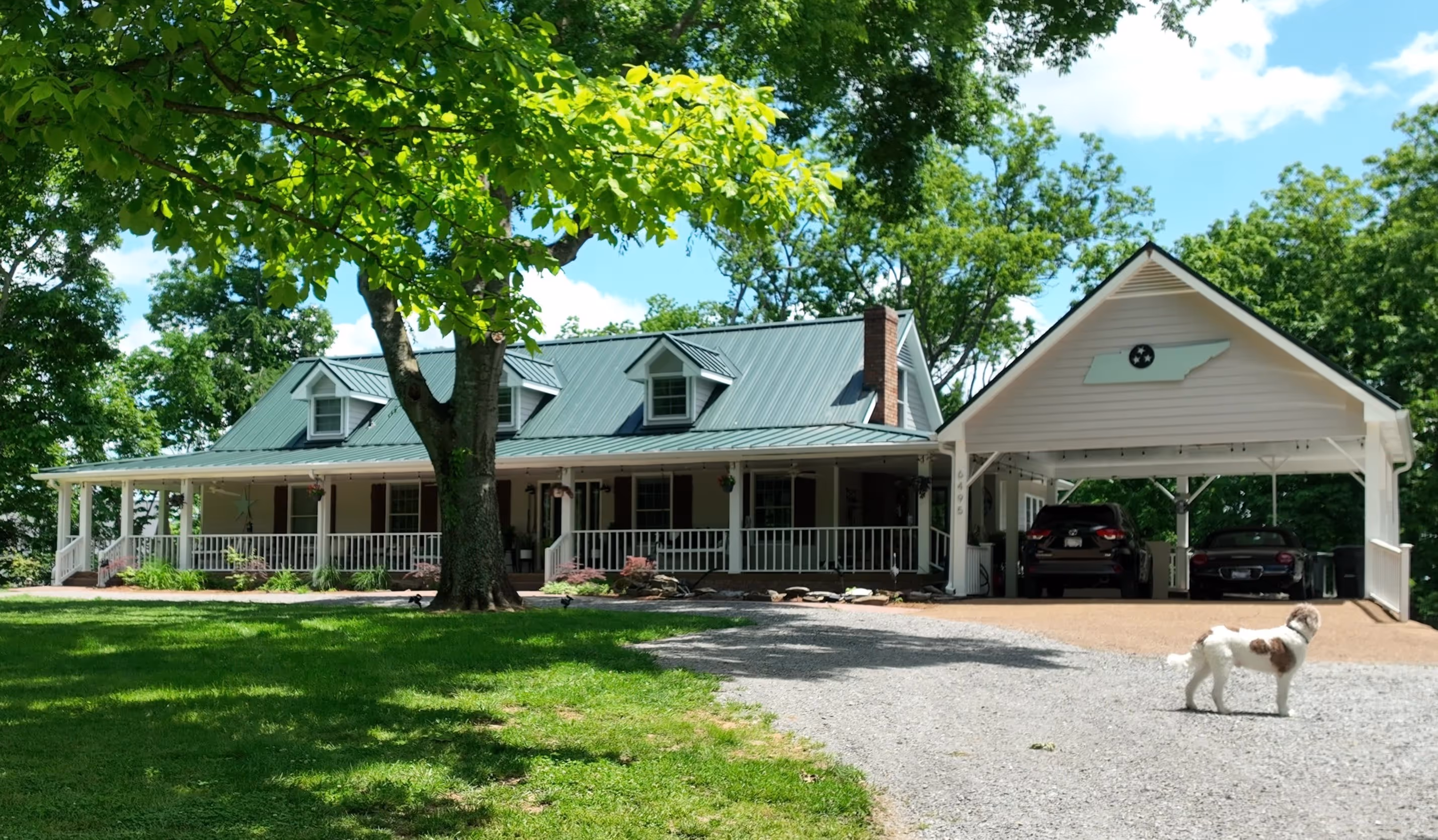 White house with green metal roof, covered porch, carport, two cars, and a dog on a gravel driveway.