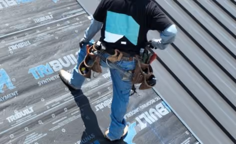 Worker wearing tool belt installing synthetic roofing underlayment on sloped roof near metal siding.