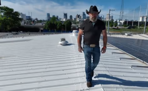 Man wearing a black cowboy hat walking on a white rooftop with a city skyline in the background.