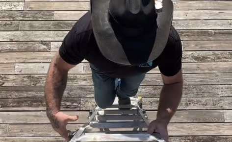 Man wearing a hat climbing a ladder on a wooden deck viewed from above.