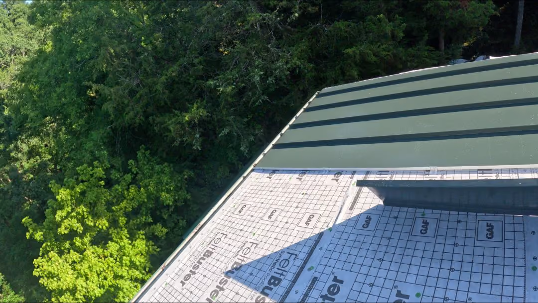 Green metal roof partially covered with roofing underlayment beside leafy green trees.